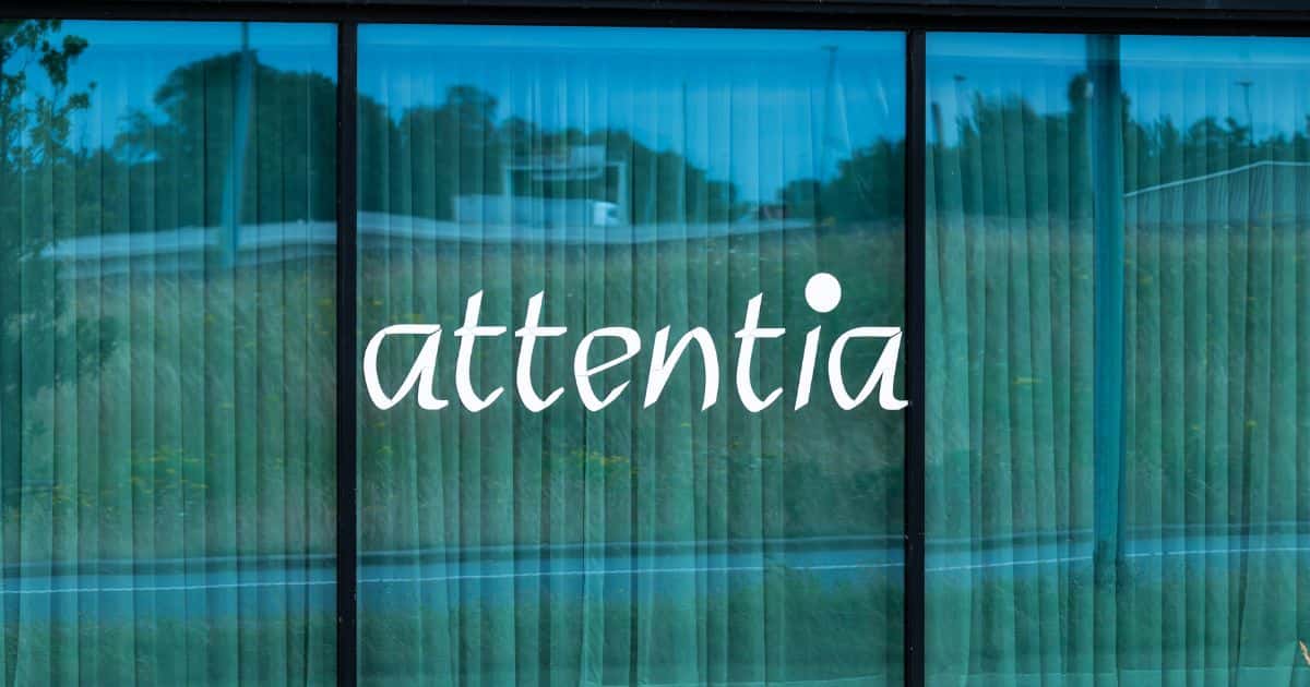 Logo van het sociaal secretariaat Attentia op een groot venster in 3 delen. In de ramen ook de reflectie van een drukke baan.