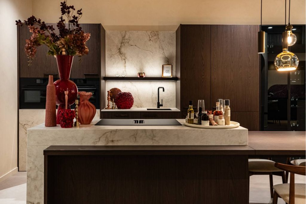 Gedetailleerde showroomweergave van een luxe keuken met donkere houtfineer kasten, een verlicht wit marmeren achterpaneel en een tweedelig kookeiland. Het eiland heeft een verhoogd marmeren kook-gedeelte en een lager, houten eettafel-gedeelte met stoelen. Opvallende rode vazen decoreren de ruimte.