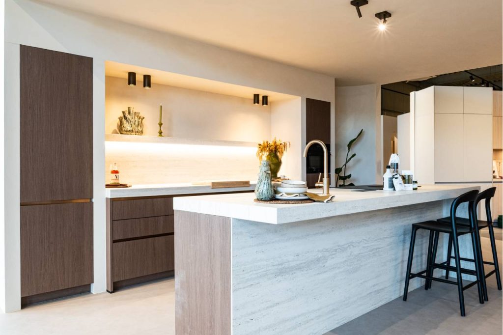 Luxe moderne showroomkeuken met donkerbruine eikenhouten kasten, een doorlopend travertijn-achtig natuurstenen kookeiland en achterwand met geïntegreerde verlichting. Twee zwarte barkrukken staan bij het eiland, en een goudkleurige kraan is zichtbaar.