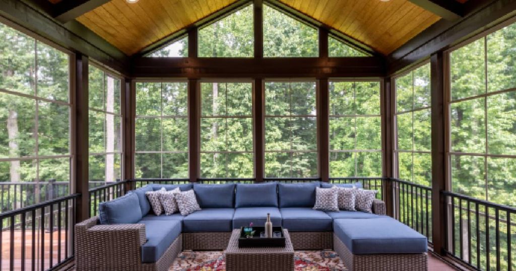 Een grote, luxe prefab veranda uit hout met grote raampartijen die uitgeven op een bos. In de prefab veranda staat een grote blauwe zetel en een salontafeltje.