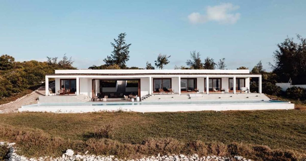 Een grote, uitgestrekte witte bungalow met een plat dak. Het gebouw heeft een breed zwembad, straalt luxe uit, en ligt ten midden van een bosrijk gebied.