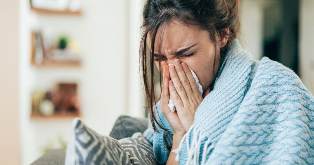 Een zieke vrouw onder een gebreid blauw dekentje snuit haar neus met twee handen. Ze zit thuis op een zetel met lichtgrijze kussens met donkergrijze strepen.