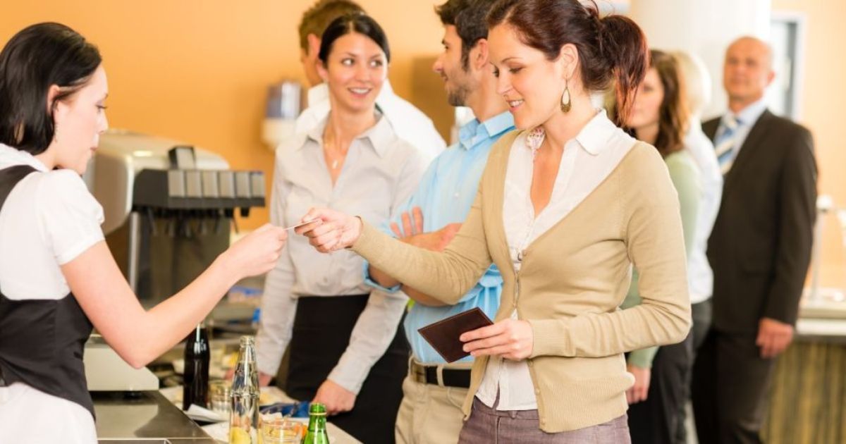 Een dame geeft haar betaalkaart met maaltijdcheques aan een dame aan de kassa. Achter haar staat een rij wachtenden met elk een plateau met eten en drinken.