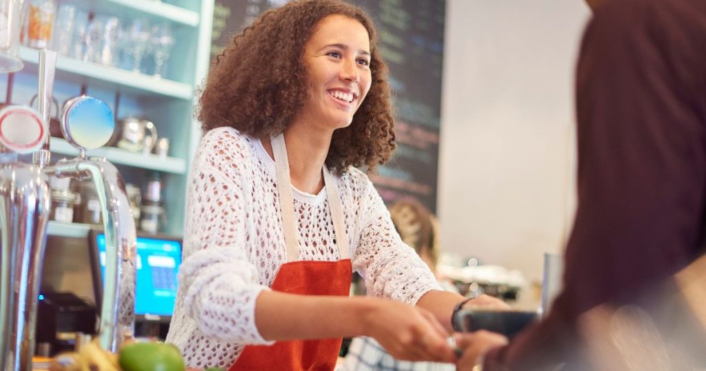 Een dame met krullend haar, een wit gehaakt vestje en een rode schort met witte bretellen bedient een klant in een horecazaak. Op de achtergrond staan een kassascherm, een krijtbord met menu in blauw, groen en rood, een kast met glazen en een machine.