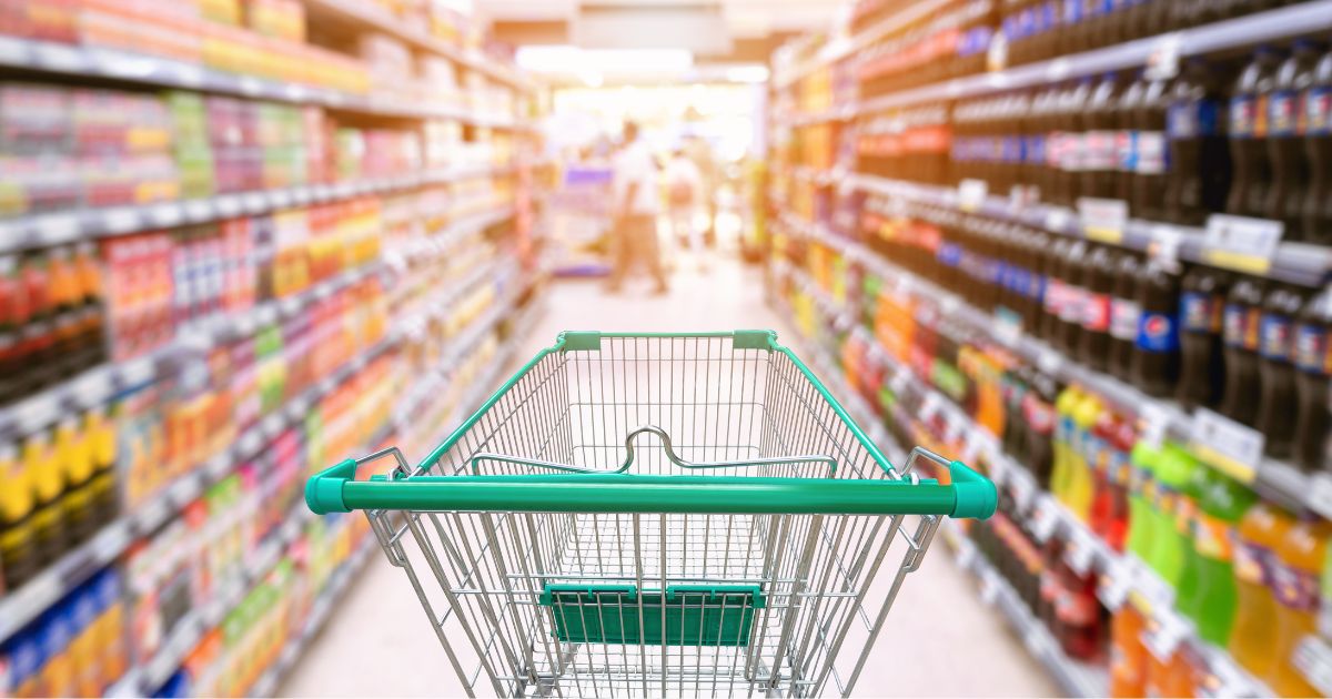 Een winkelkarretje met groene details tussen 2 rijen met drank in een supermarkt.