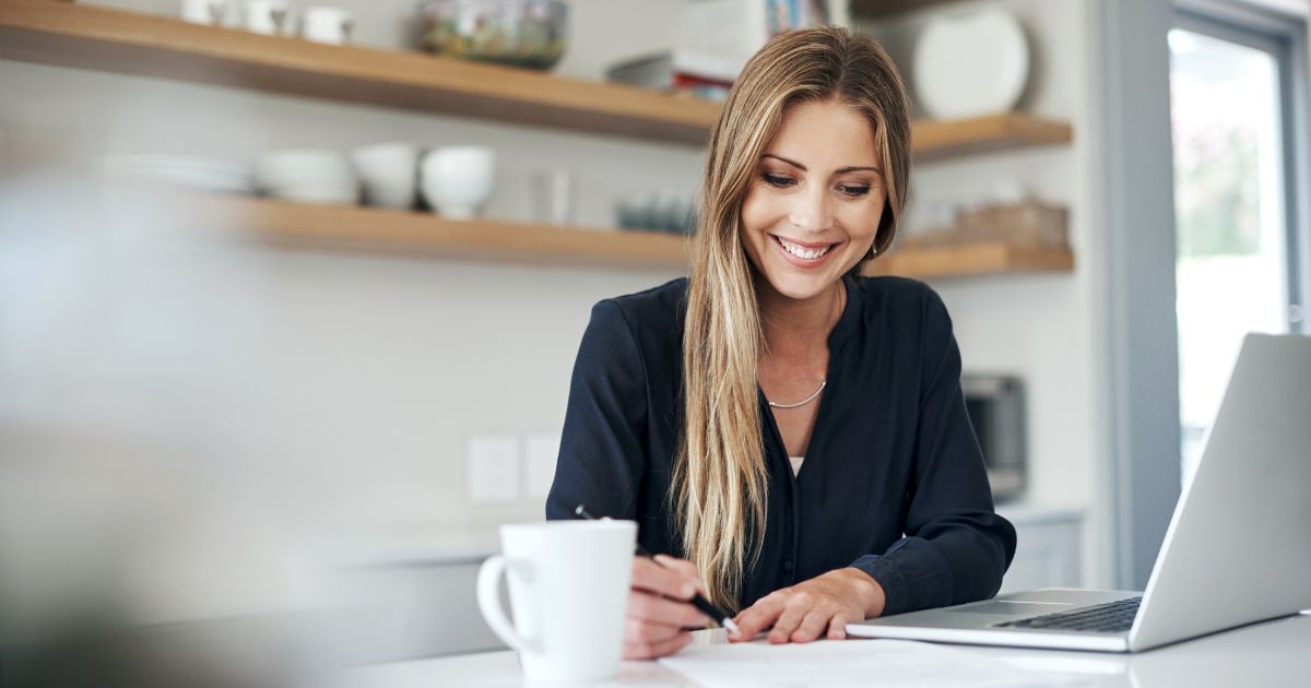 Een dame in zwarte blazer zit achter een laptop in een keuken. Voor haar staat een witte tas.
