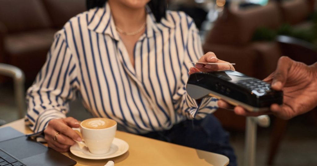 Een dame in gestreept hemd betaalt contactloos via een betaalterminal. Voor haar op tafel staat een kop koffie.