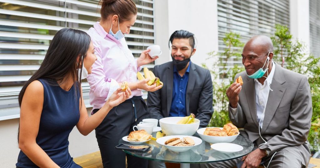 Vier collega's eten samen aan een glazen tafeltje in open lucht.