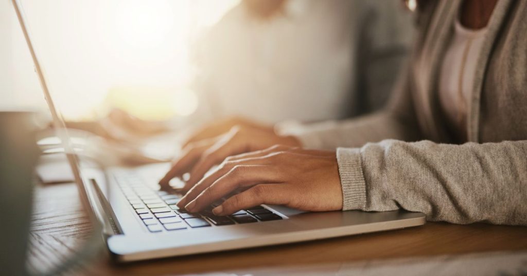 Twee handen in grijze mouwen typen op een grijze laptop.