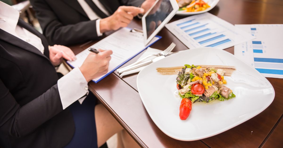 Twee personen zitten aan tafel: één persoon houdt een tablet vast en wijst met een vinger naar het scherm. Terwijl de andere persoon notities maakt op een blad. Op de tafel staat ook een bord met een slaatje.