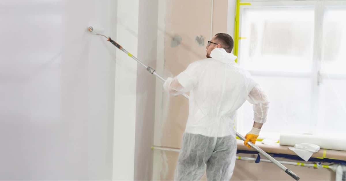 Een man draagt transparante, beschermende kledij terwijl hij met een roller de muur wit verft. Ook de rest van de kamer rondom hem is afgeplakt met tape en transparante plastiek.