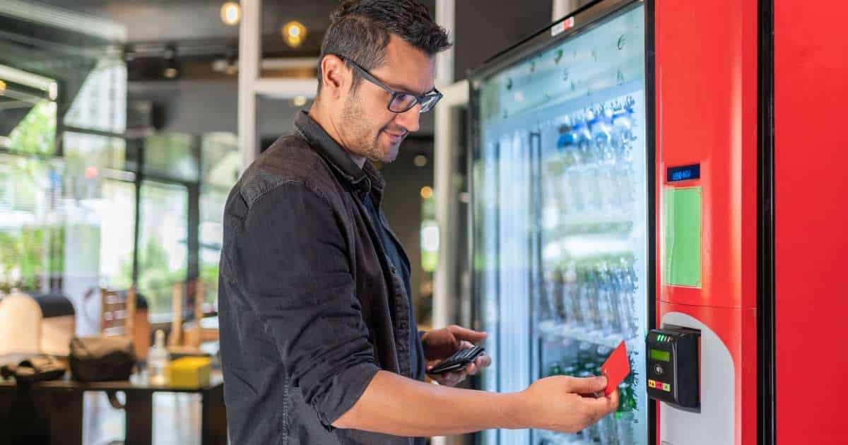 Een man gebruikt een rode bankkaart om contactloos te betalen bij een rode drankautomaat. Hij draagt een bril, zwartgrijs vest en een baardje.
