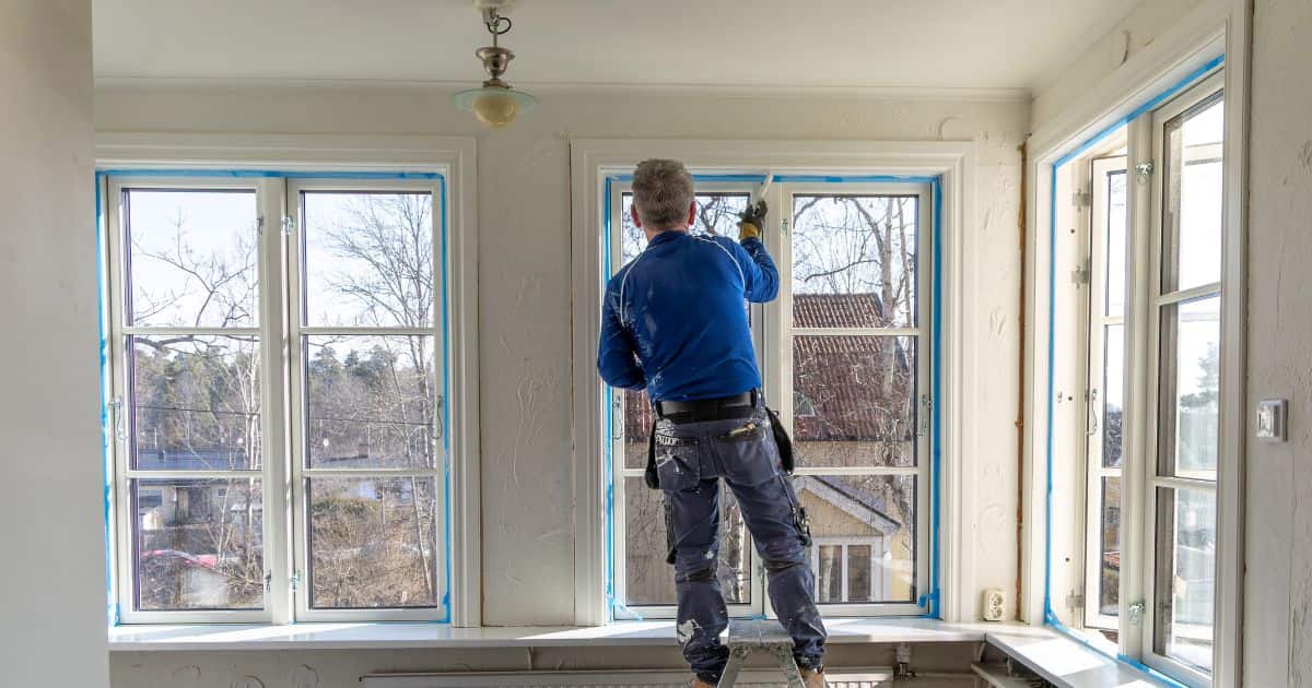 Een man met blauw t-shirt heeft de wanden naast de ramen professioneel afgetapet, zodat hij makkelijk de raamkozijnen kan wit schilderen.