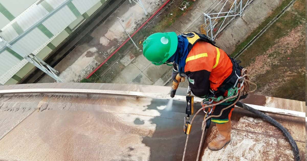 Een man met groene helm en oranje veiligheidstrui richt het zandstraalpistool op een stalen of metalen oppervlak. Rondom de gereinigde plek spoelt het roest weg. De man werkt op hoogte: hij draagt een veiligheidsgordel waaraan musketons en koorden zijn vastgemaakt.