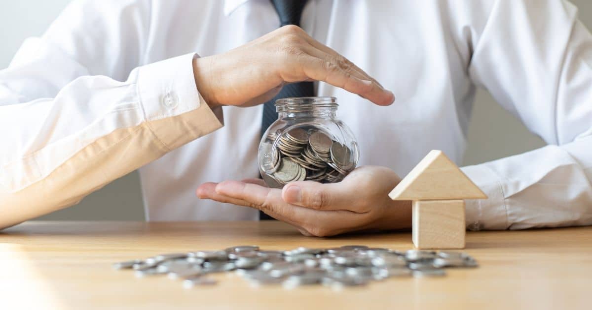 Een man in wit hemd met een zwarte das houdt een glazen potje vast. In het potje zitten muntstukken. Voor hem, op een lichthouten tafel, liggen meer muntjes. Naast zijn linkerhand staan 2 op elkaar gestapelde houten blokken: een kubus en een driehoek.