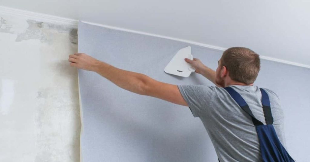 Een man in grijs t-shirt en blauwe overall strijkt een strook behangpapier vast en glad op een muur. Voor een perfect resultaat houdt hij daarbij het uiteinde van het papier vast.