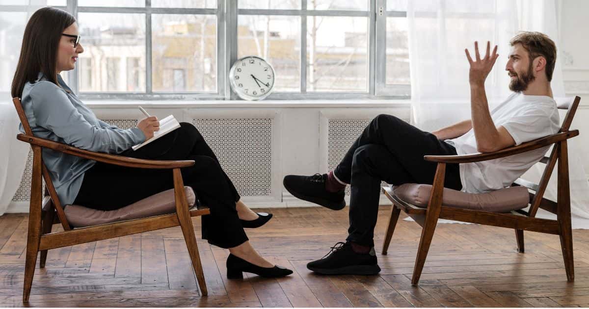 Een vrouwelijke psycholoog en haar volwassen, mannelijke patiënt in een witte ruimte met houten vloer. De psychologe zit recht tegenover haar patiënt en heeft een klembord met pen vast. Op de achtergrond een klok.