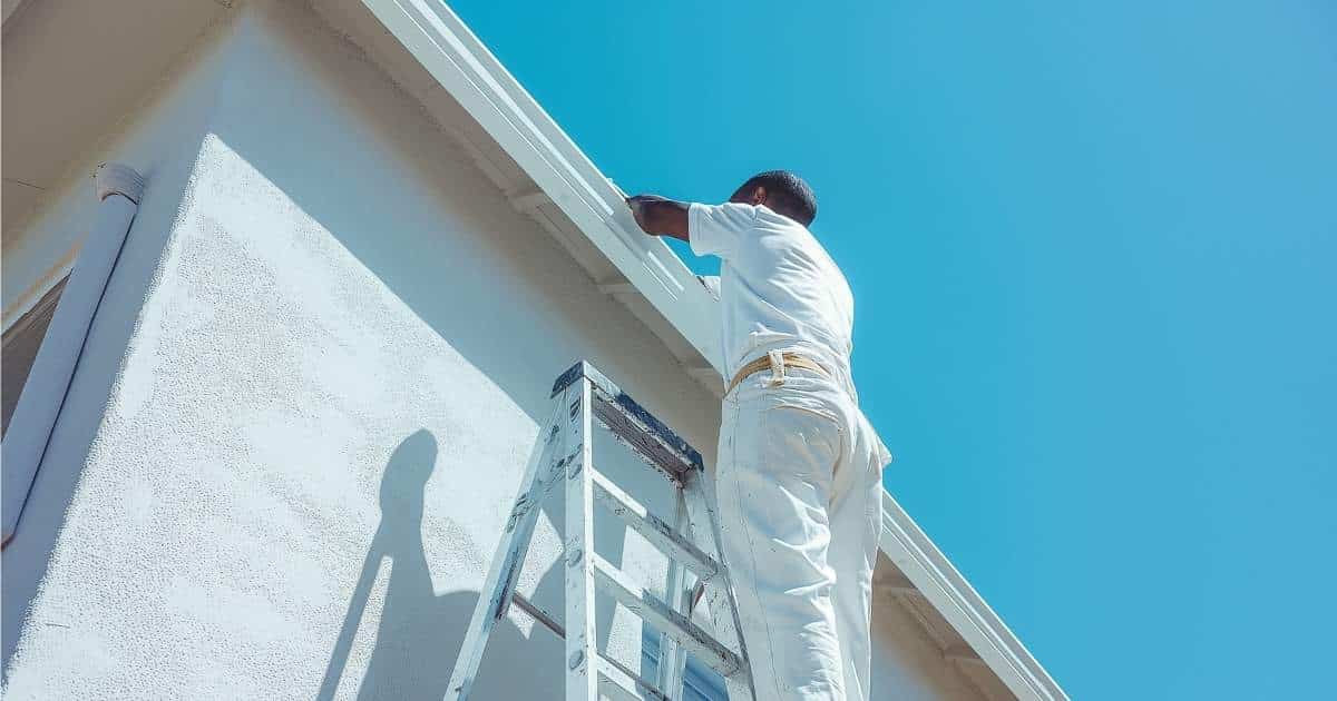 Een man in witte broek en wit t-shirt werkt aan een dakrand. Hij staat op een ladder die tegen een witte crepi gevel staat.