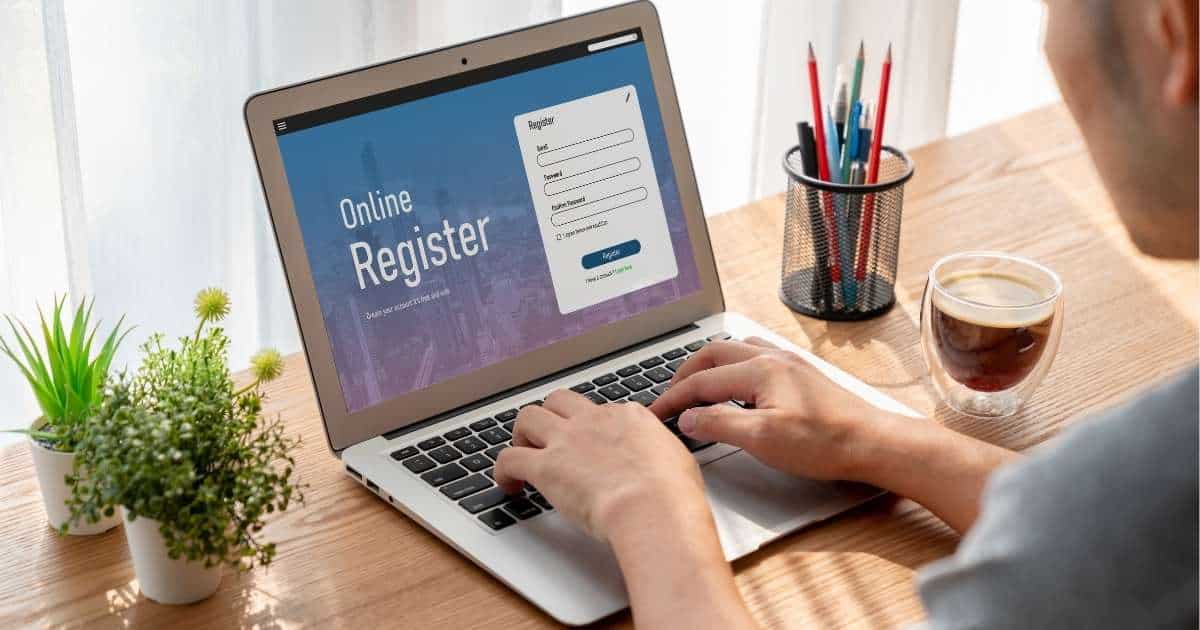 Een man met grijs t-shirt zit achter zijn laptop, die op een houten tafel staat. Hij meldt zich aan via het online tijdsregistratiesysteem van zijn werkgever. Links van zijn laptop staan twee kamerplantjes in witte potjes. Rechts van de laptop staat een pennenhouder met schrijfgerei en een doorzichtige tas met koffie zonder melk.