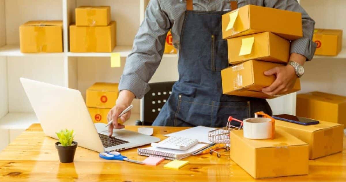 Een webshopuitbater overloopt de bestellingen die klanten bij hem plaatsten via zijn online betaalsysteem. Hij draagt een jeansoveral over een grijs hemd en heeft enkele pakketjes onder zijn arm.