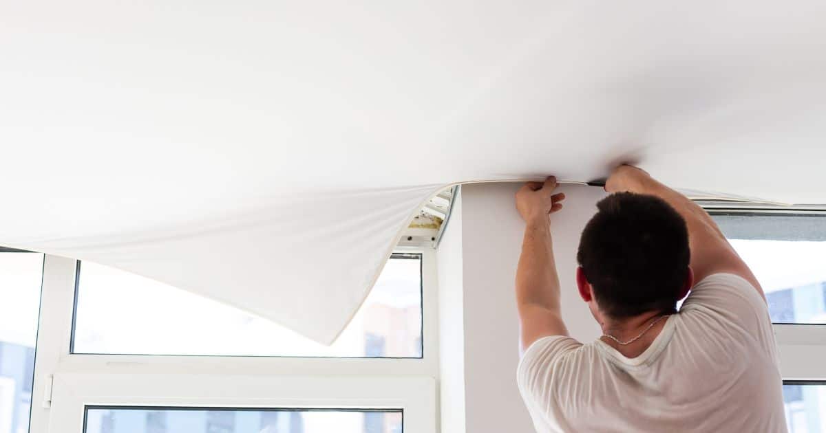 Een vakspecialist plaatst een koud polyester spanplafond in een woonkamer. Hij spant het doek tot in de profielen die langs de wand en ramen in de ruimte lopen.