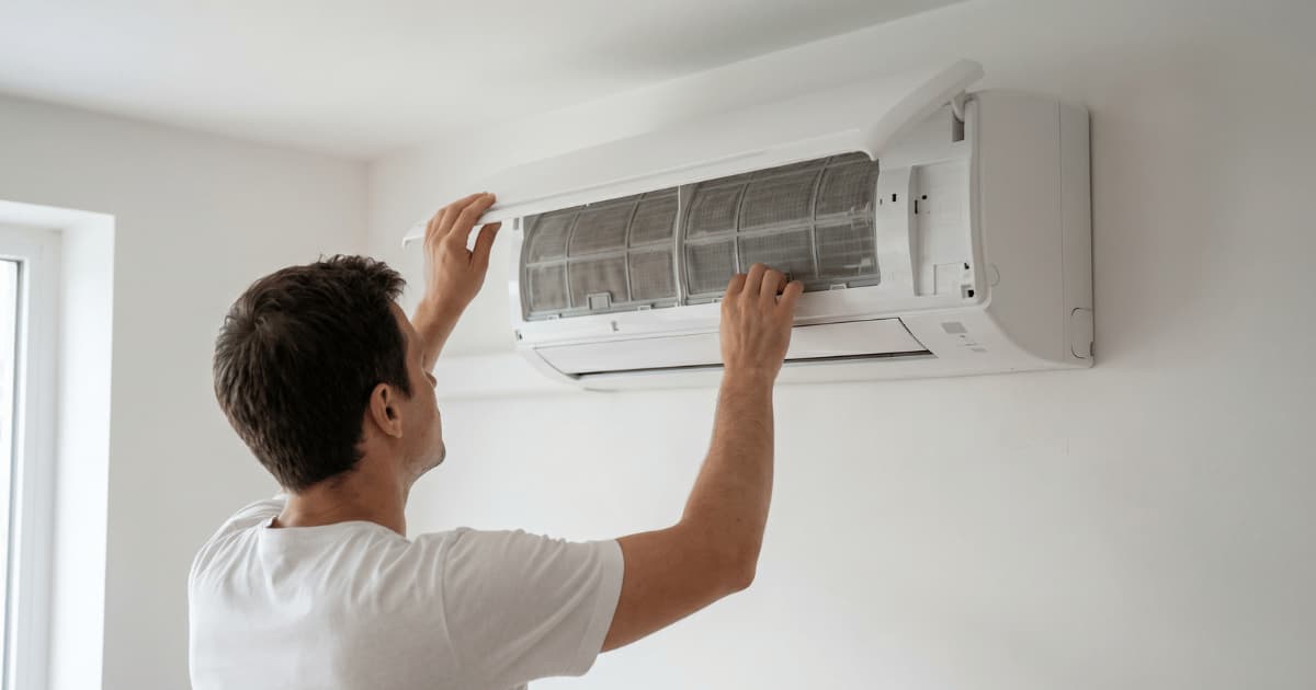 Man in wit t-shirt opent met beide handen het frontpaneel van een witte airco binnenunit aan de muur waardoor de stoffilters zichtbaar worden.