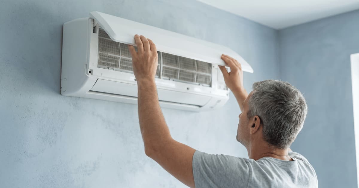 Een man reikt omhoog en opent het frontpaneel van een witte airco-binnenunit aan de muur waardoor de filters zichtbaar worden.