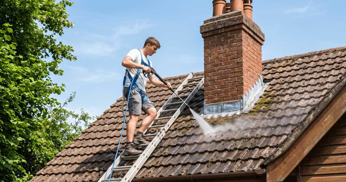 Een jonge man in een wit t-shirt en blauwe veiligheidstuig staat op een aluminium ladder tegen een hellend dak en spuit met een hogedrukreiniger op verweerde bruine betonnen dakpannen nabij een bakstenen schoorsteen met zichtbare mosgroei en loodslabben terwijl groene boomtakken het beeld links omkaderen.