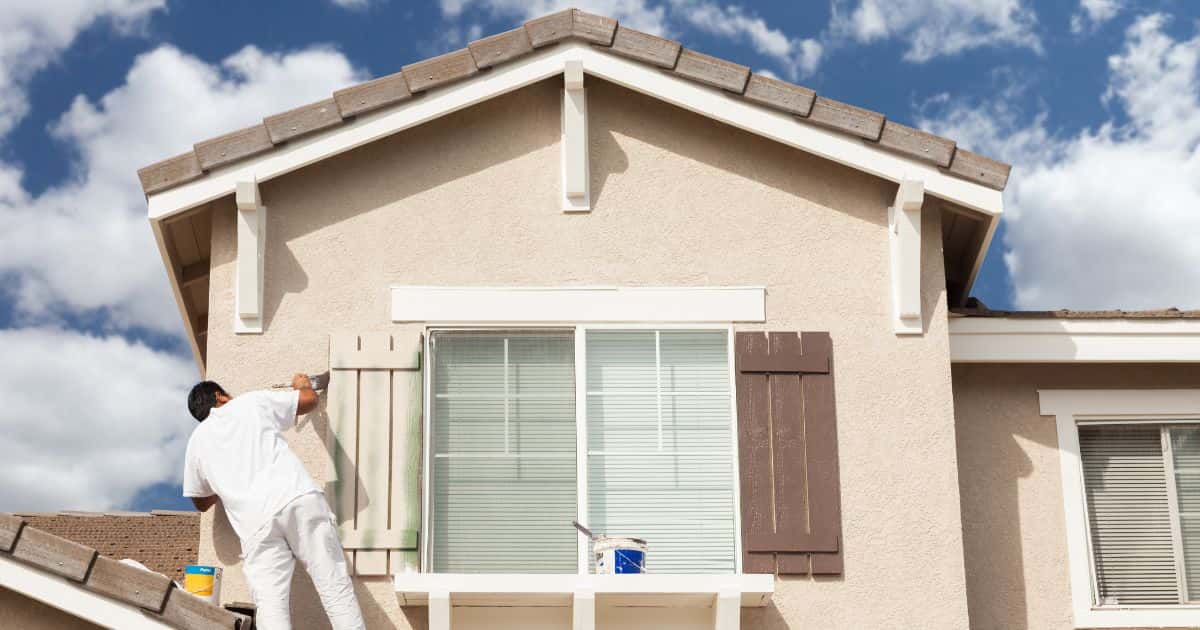 Een man in wit t-shirt en broek )schildert de luiken van de ramen. Ze voegen een leuk detail toe aan de rest van de woning.