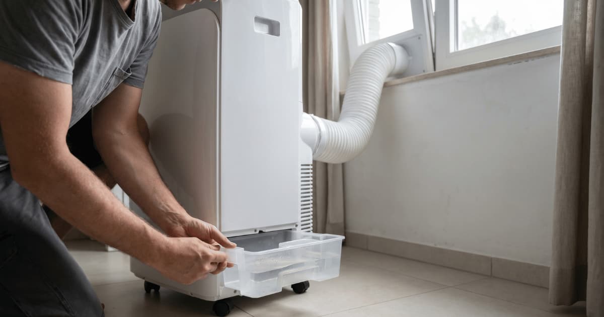 Een man in grijs t-shirt hurkt naast een witte mobiele airco op wieltjes en schuift met beide handen de doorzichtige condensbak met water uit de onderkant van het toestel terwijl de witte afvoerslang naar het raam loopt.