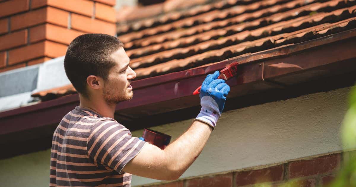 Een man met kort haar en streepjes t-shirt verft een dakgoot rood/bordeaux. In zijn rechterhand houdt hij de verfborstel vast, in zijn linkerhand de verfpot.