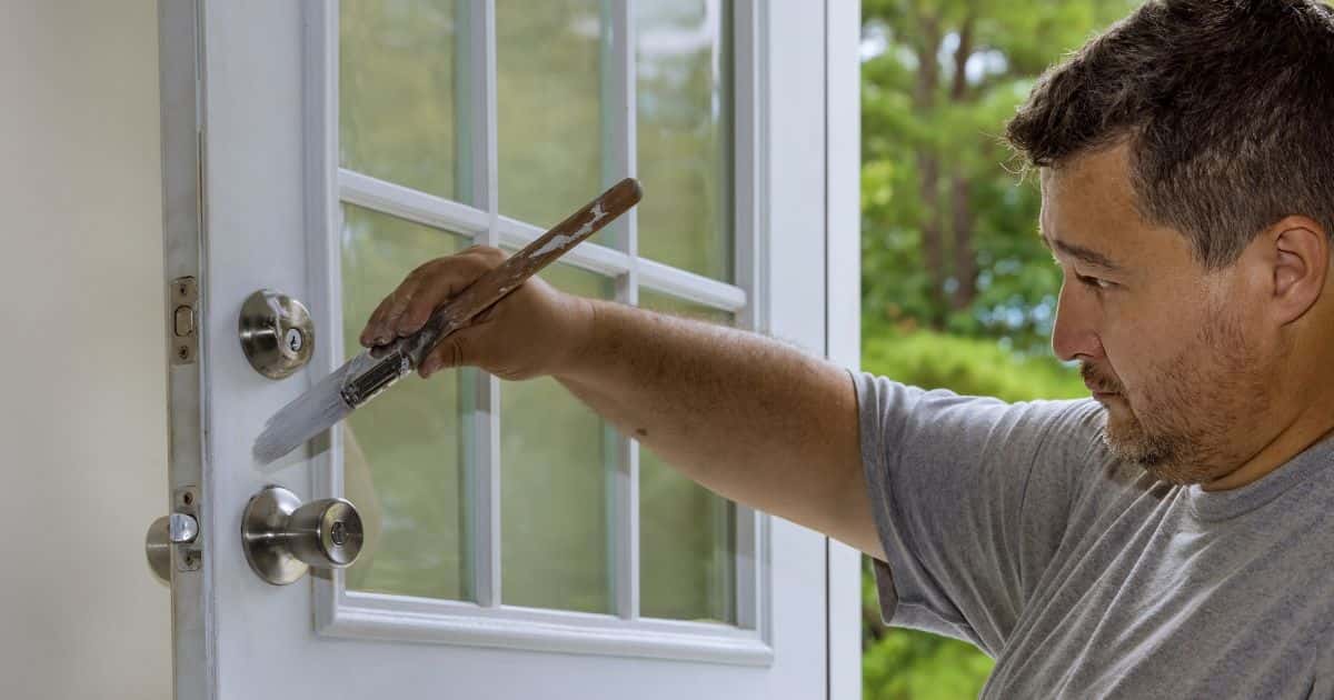 Een man met stoppelbaard focust zich op het wit verven van een buitendeur in zijn woning. Hij gebruikt daarvoor een fijnere verfborstel.