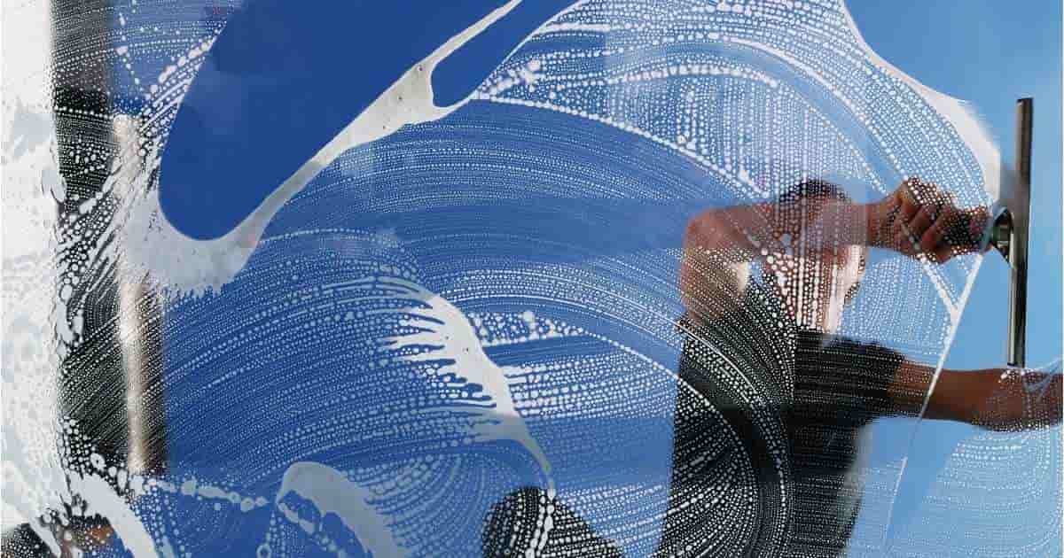 Een raam met zeepsop. Daarachter is een man in zwart t-shirt zichtbaar. Hij verwijdert zeepsop van een raam met een zwarte trekker.