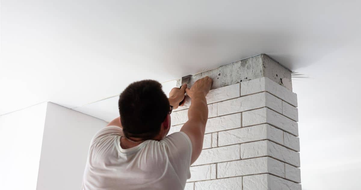 Een vakspecialist plaatst een spanplafond in een keuken. Hij werkt de hoeken van het spanplafond af rond een betegelde zuil in de ruimte.