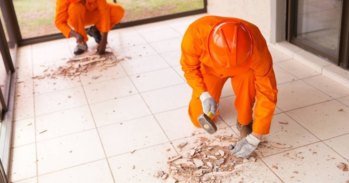 Twee vloerders in oranje werkkledij, oranje helmen en werkhandschoenen zijn bezig met het uitbreken van een tegelvloer.