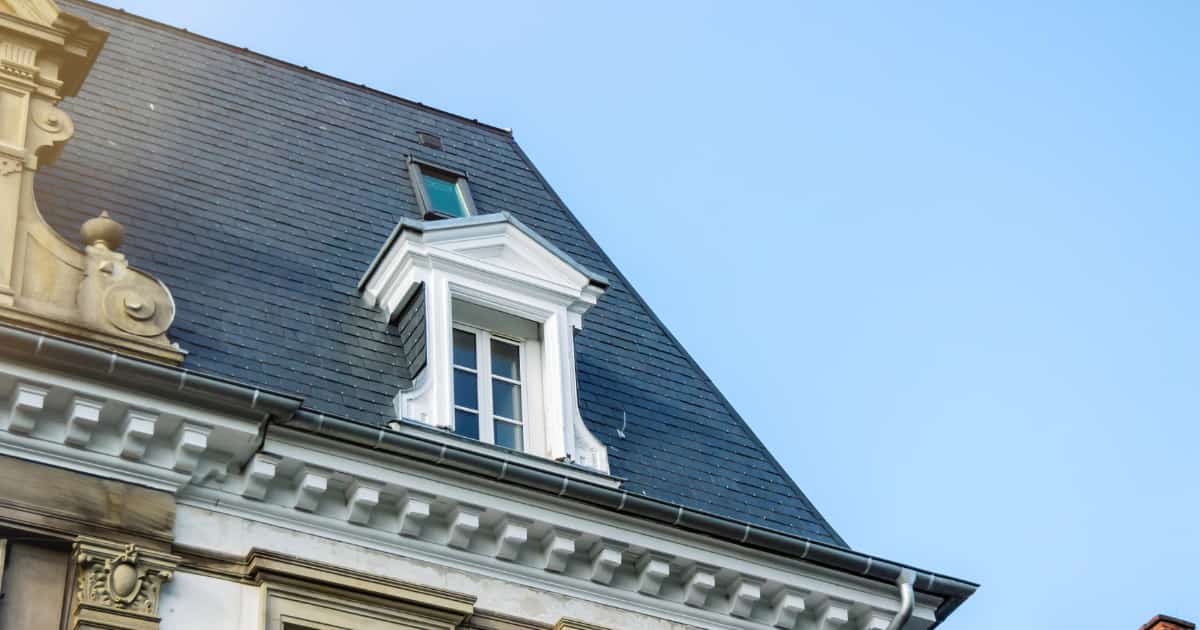 Een steil mansardedak bedekt met donkergrijze natuurleien op een klassiek herenhuis met een sierlijke witte dakkapel met driehoekig fronton en een klein dakraam hoger op het dakvlak boven een ornamentale kroonlijst.