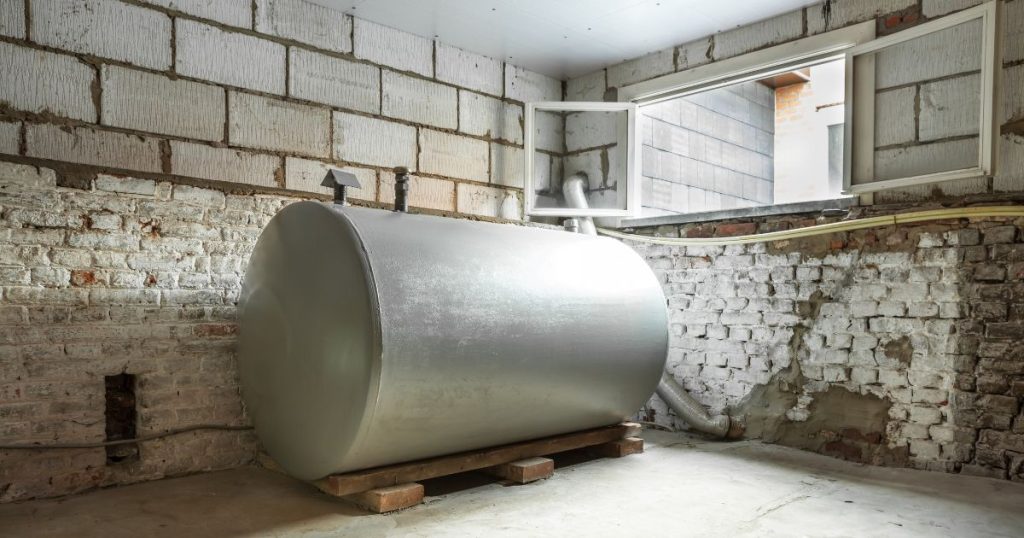 Een oude, metalen, bovengrondse mazouttank in een onafgewerkte kelder. De mazouttank staat op houten planken. De kelder beschikt over een raam en heeft witte, bakstenen muren en een betonnen vloer.