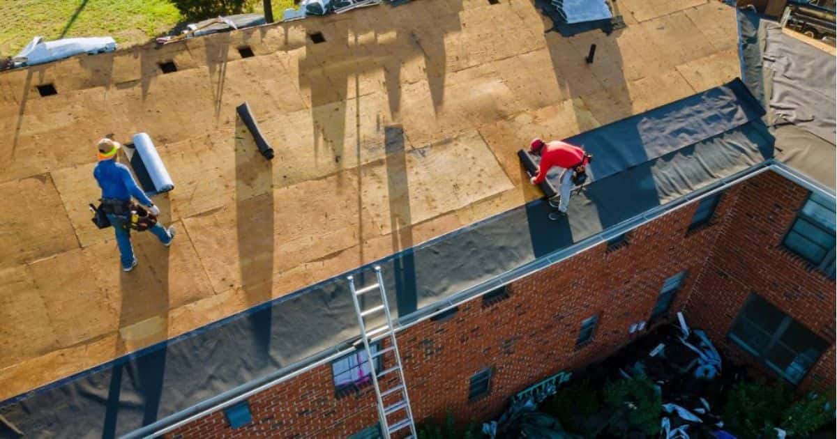 Twee werkmannen zijn bezig op het dak van een stenen gebouw. De man in blauwe werkkledij stapt op de houten panelen richting een rol folie. De man met blauwe jeans en rood t-shirt is bezig met het uitrollen van een zwarte rol folie.