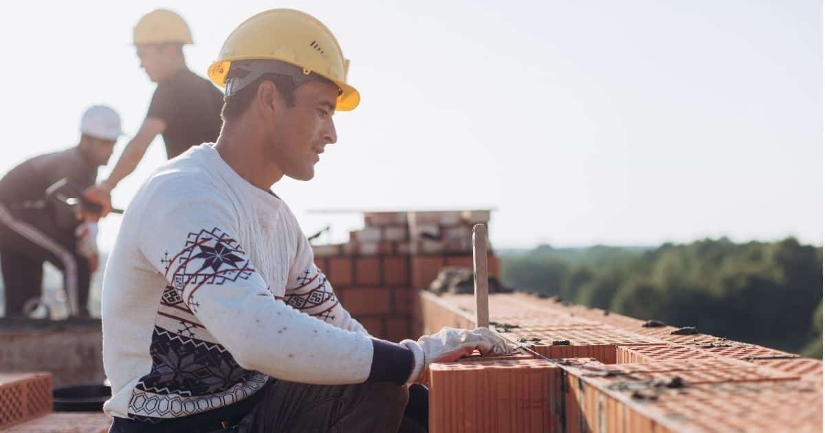 Een metser metselt snelbouwstenen in de ruwbouwfase van een aanbouw- of nieuwbouwproject. Hij draagt een witte trui met kerstmotief, witte werkhandschoenen en een gele veiligheidshelm.