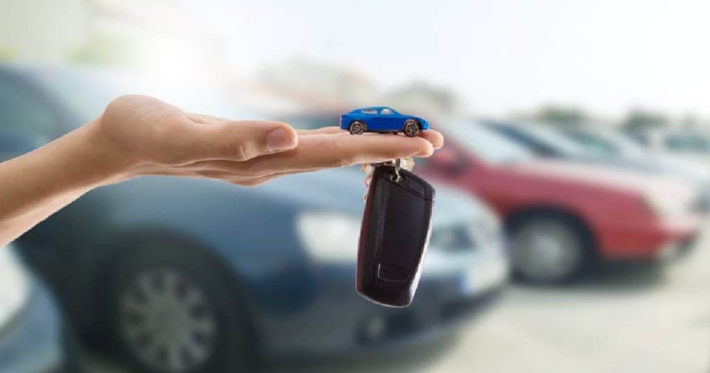 Een hand met opgeheven handpalm. Op de vingers ligt een blauw miniatuurautootje. Aan de ringvinger hangt een zwarte autosleutel. Op de achtergrond geparkeerde wagens op een parking.
