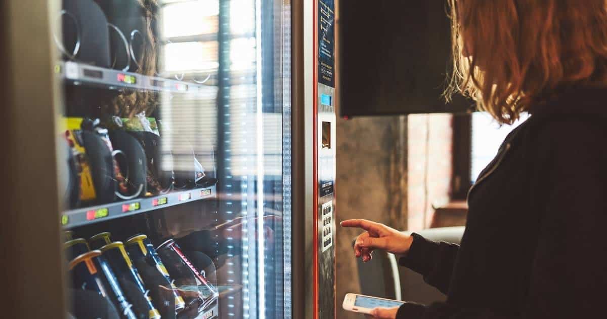 Een dame drukt een code in op het bedieningspaneel van een drankenautomaat met dranken en snacks.