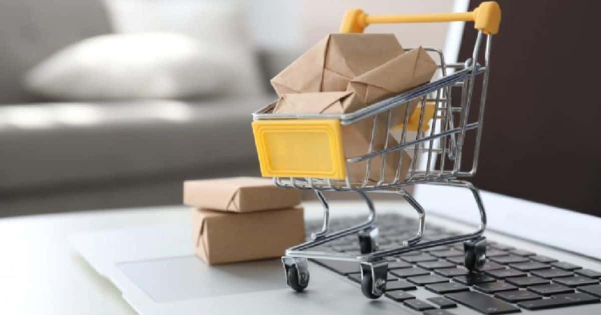 Een mini winkelwagentje gevuld met pakketjes die klanten bestelden via een online betaalsysteem van een webshop staat op een laptop. Naast het kleine winkelwagentje met een gele stootrand staat een stapeltje met twee kleine pakketjes.