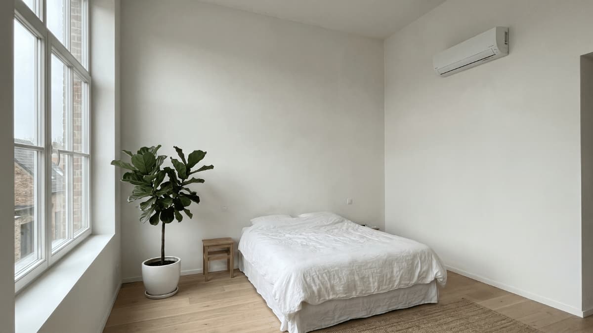 Een minimalistische slaapkamer met hoge witte muren en lichte eiken vloer waar een witte split-airco binnenunit hoog aan de rechtermuur hangt terwijl een grote vijgenboomplant naast het raam staat en door de hoge ramen bakstenen daken zichtbaar zijn onder daglicht.