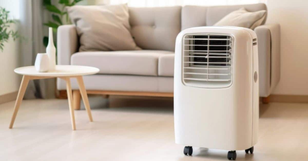 Een witte mobiele airco staat in het midden van een woonkamer. Achter de mobiele airco staat een beige bank en een kleine koffietafel.