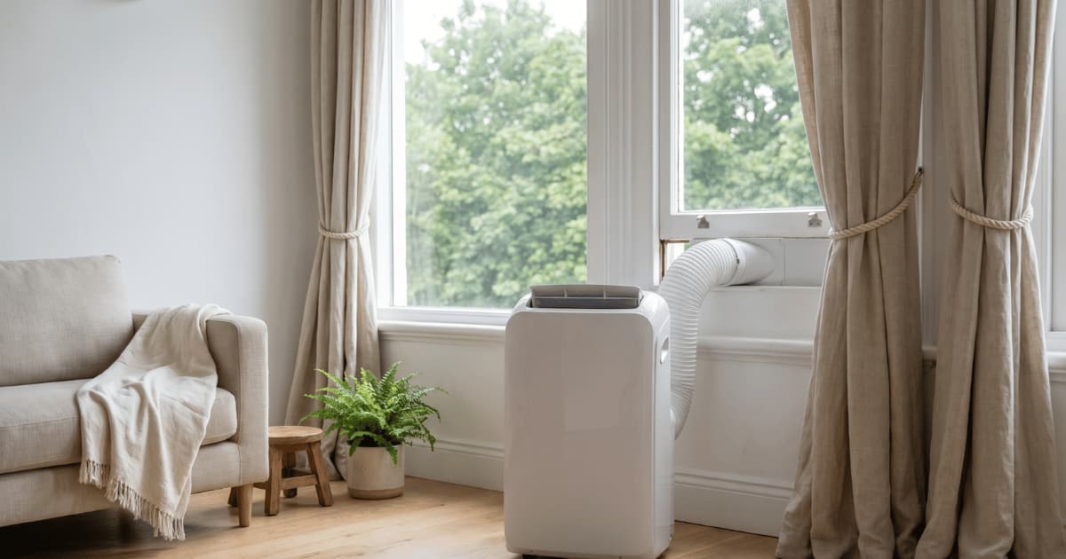 Witte mobiele airco met afvoerslang door een open raam in een lichte woonkamer met linnen gordijnen.
