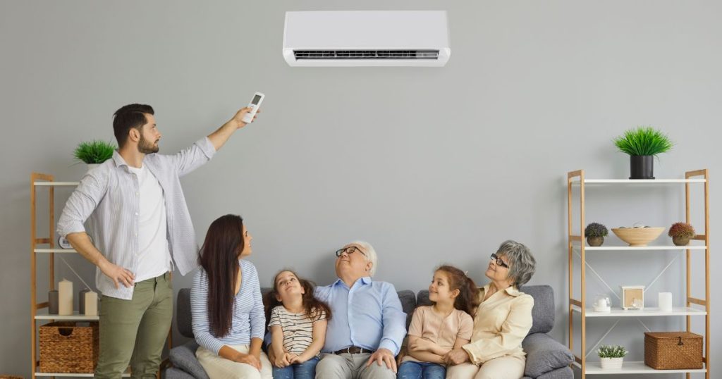 Een man (links) gebruikt een afstandsbediening om de binnenunit van een multi-split unit airco systeem te bedienen: een toestel dat je het hele jaar door controle geeft over het binnenklimaat in huis. Op de zetel onder de airconditioner unit zitten, van links naar rechts, een vrouw, een jong meisje, een oudere man, een jong meisje en een oudere dame.