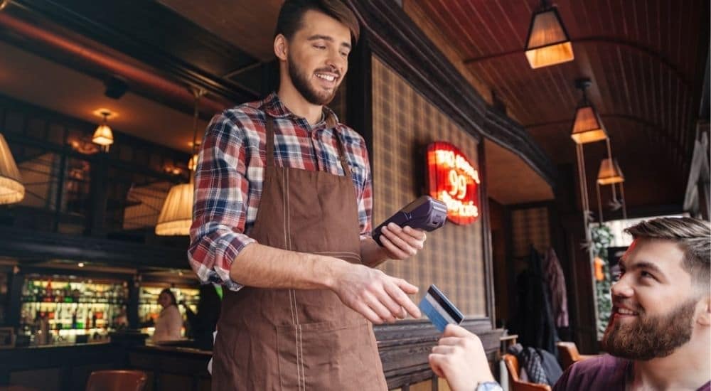 Een ober in een bruine schort helpt een klant bij het afrekenen met een mobiele betaalterminal.