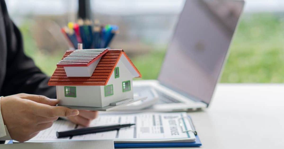 Persoon houdt een modelhuisje vast boven een bureau met documenten en een laptop.