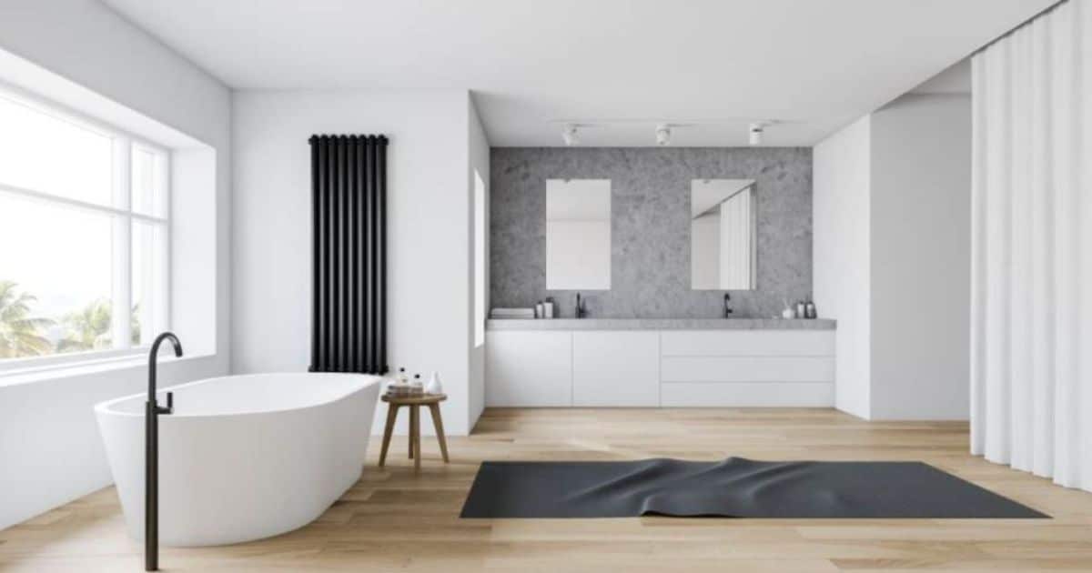 Moderne badkamer met een vrijstaand wit bad, dubbele wastafel met grijze marmeren wand en spiegels, zwarte radiator en lichte houten vloer.