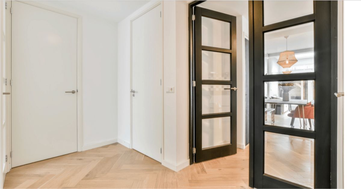 Een hal met twee witte deuren en een derde, openstaande deur. Deze deur heeft een zwarte omlijsting met glas erin. De deur leidt naar een woonkamer met een eettafel en stoelen
